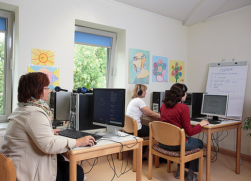 Gruppenraum mit PC-Ausstattung für Cogpack: An vier PCs sitzen Personen mit Kopfhörern, die konzentriert arbeiten. Im Hintergrund ist ein Flipchart zu sehen.