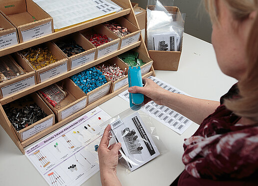 Eine Person sitzt an einem Tisch. Auf dem Tisch sind viele kleine Schütten zu sehen, die unterschiedliche elektronische Bauteile enthalten. Die Person heftet gerade eine durchsichtige Tüte zu, die mit diesen Teilen befüllt wurde.