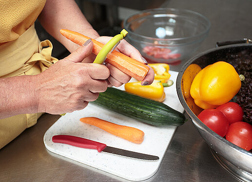 Gemüseschälen: Auf dem Bild sind die Hände und Arme einer Person zu sehen, die gerade eine Karotte schält. Auf dem weißen Schneidbrett sind ein scharfes Messer, eine weitere Karotte, eine ungeschälte Gurke und eine geteilte gelbe Paprika zu sehen. In einer Schüssel, die am rechten Bildrand erkennbar ist, liegen noch eine weitere gelbe Paprika und einige Tomaten.