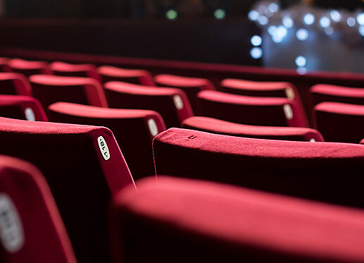 Es sind mehrere Reihen mit rotem Stoff bezogene Kinosessel zu sehen. Die Kinoleinwand ist im Hintergrund zu erahnen.