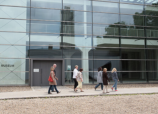 Auf dem Foto spaziert eine kleine Gruppe von sieben Personen an einer überdimensionalen Glasfront vorbei. Links ist die Aufschrift „Museum“ zu lesen.