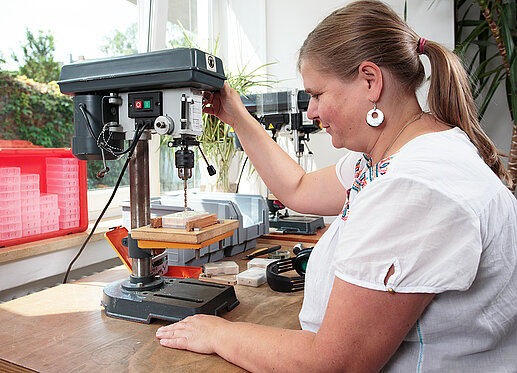 Bohrmaschine: Eine Frau sitzt an einem Arbeitstisch und bedient eine Tischbohrmaschine.