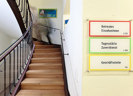 Treppenaufgang des Standorts an der Esplanade 1: Im Zentrum des Bildes ist eine breite, gewundene Holztreppe zu sehen. Rechts im Bild sind Hinweisschilder zu erkennen, auf denen „Betreutes Einzelwohnen“, „Tagesstätte/Zuverdienst“ und „Geschäftsstelle“ steht.