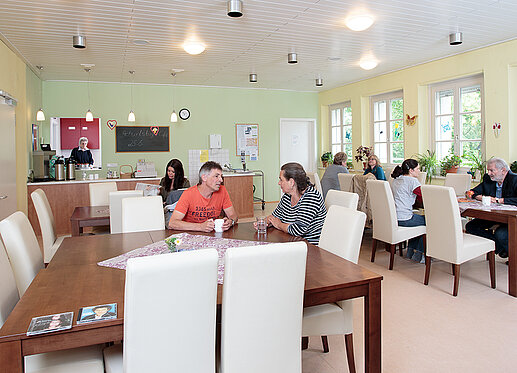 Cafeteria Esplanade: In einem großen, in hellem Gelb und lindgrün gehaltenen Raum, sitzen an verschiedenen Tischen verteilt Besucher und Besucherinnen der Cafeteria. Einige unterhalten sich oder lesen etwas. Im Hintergrund ist eine Theke zu sehen, auf der Kaffeekannen stehen. An der Wand hängen Programmhinweise.