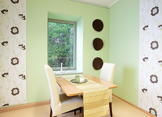Beratungsraum: Auf dem Bild ist ein Tisch mit zwei gegenüberliegenden hochlehnigen Stühlen zu sehen. Der Raum strahlt Ruhe aus. Die Wand ist in lindgrün gehalten; unterbrochen von zwei Tapetenbahnen, die ein dezentes Blumenmuster haben.