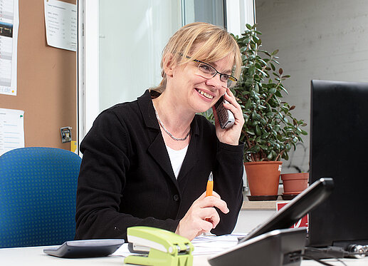 Telefonat: Eine Frau sitzt mit Kugelschreiber in der Hand an einem Schreibtisch. Sie telefoniert.