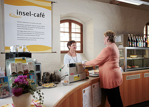 insel-café in der Stadtbücherei: An einer geschwungenen Theke stehen sich die Servicekraft und eine Kundin, die gerade eine Tasse Kaffee entgegennimmt, gegenüber. An der dicken Wand im Hintergrund hängt ein großes Plakat mit dem Hinweis auf das insel-café. Auf der Theke stehen die Teesorten zur Auswahl. Hinten rechts erkennt man die Tortenauslage und die Getränkeauswahl.