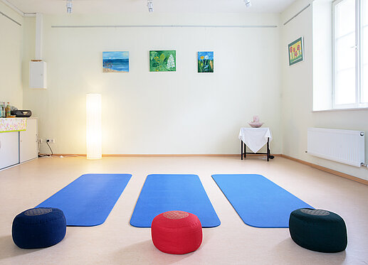 Yoga-Raum: Es ist ein Raum mit hellen Wänden zu sehen. Auf dem Boden liegen nebeneinander drei blaue Gymnastikmatten und je ein verschiedenfarbiges Sitzkissen. An der Wand hängen einige ruhige Aquarelle, vom seitlichen Fenster strömt Licht in den Raum, der dadurch eine helle, freundliche Ausstrahlung bekommt.