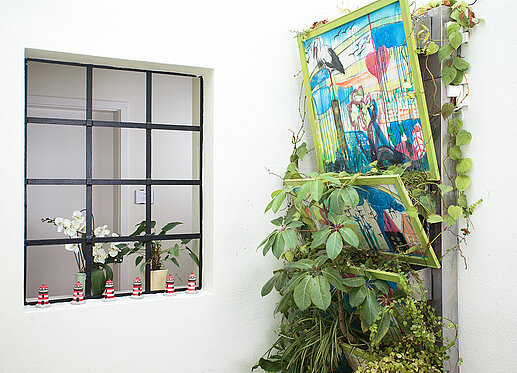 Vertikaler Garten: Links ist ein Treppenhausfenster zu sehen, das den Blick auf den dahinterliegenden Gang erlaubt. Rechts ist an der Wand eine Stahlkonstruktion zu sehen, in deren aufgefächerten Blumenkästen grüne Zimmerpflanzen wuchern.